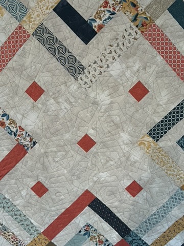 Close-up of the Bakers Dozen Quilt showing coral diamond accents, patterned strip piecing, and fine angular quilting lines